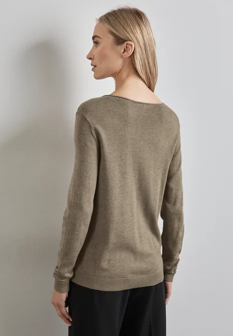 Frau mit beigem Pullover steht seitlich vor einer grauen Wand.
